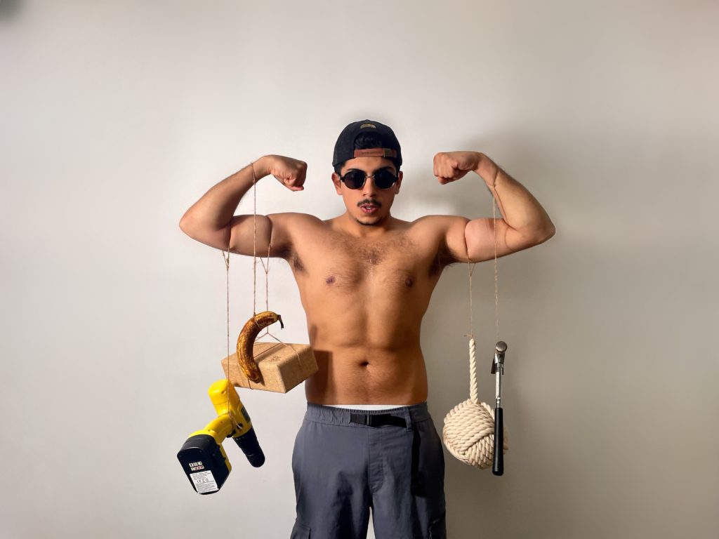 Hamza Ali, a brown-skinned man, stands topless wearing a backwards cap and sunglasses in a bodybuilder pose. He has a drill, banana, hammer, brick and ball of rope hanging from his arms using string.